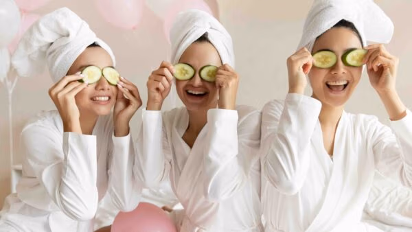 Happy Young Women Wear White Bathrobes Towels On Head Make Cucumber Facial Skin Care Mask On Eyes Laughing Relaxing Together, Smiling Having Fun On Spa Beauty Salon Party